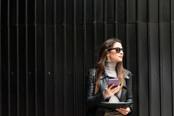 Woman in sunglasses and leather jacket holding a phone and tablet, standing in front of a black wall.