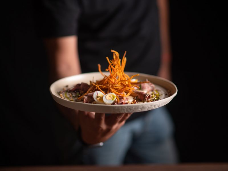 A person holds a plate of gourmet food, topped with crispy strips and sliced boiled quail eggs.