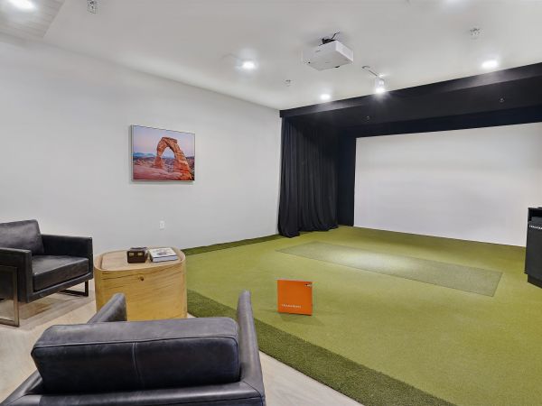Modern indoor golf simulator room with green turf, two black chairs, and a large projection screen at The Hendrey.