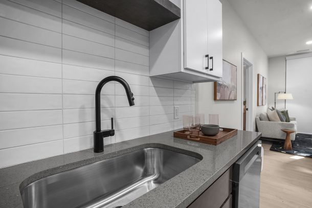 Modern kitchen sink with black faucet, gray countertop, and a tray with glasses; living area visible in background at The Hendrey.