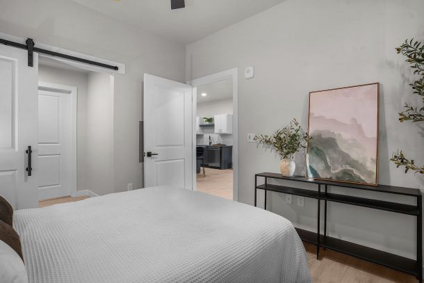 Modern bedroom with a white bedspread, black console table, framed art, and view into a kitchen area.
