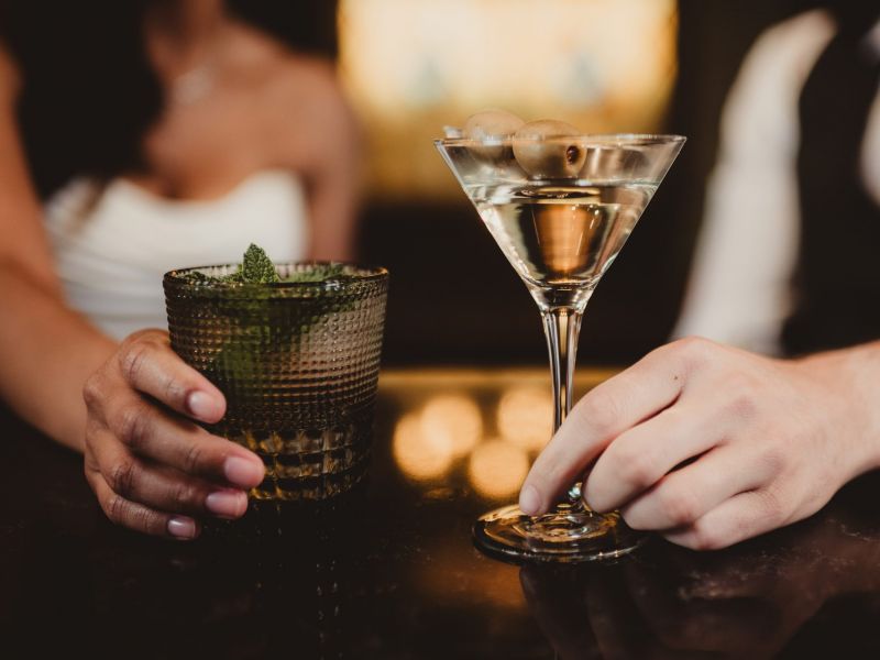 A woman and man holding cocktails at a bar, one with a mint garnish and one with olives in a martini glass.