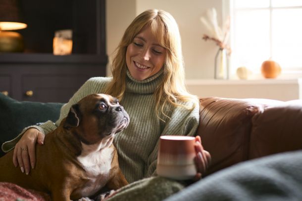 A woman in a sweater smiles while holding a mug and sitting with her dog on a couch.