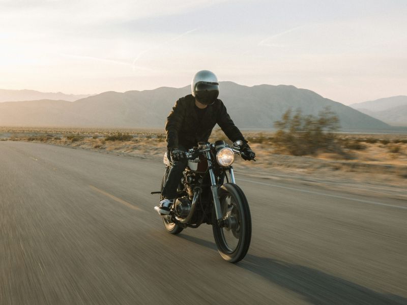A person wearing a helmet rides a motorcycle down a deserted road with mountains in the background.