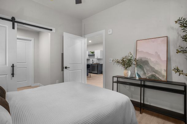 Modern bedroom with a white bedspread, black console table, framed art, and view into a kitchen area.